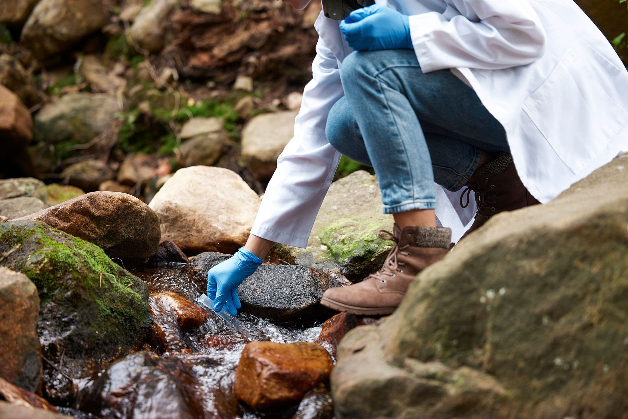 Environmental consultant in boots and gloves collecting water samples from a rocky streambed, representing site assessments for regulatory compliance.